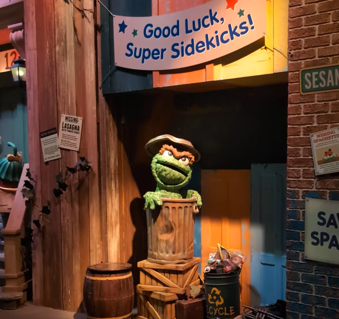 Oscar the Grouch peeks out from a trash can in a colourful set. Sign reads "Good Luck, Super Sidekicks!"