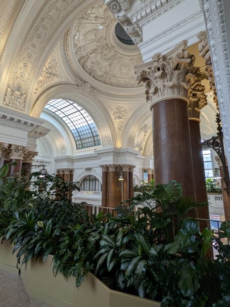 Interior of a grand hall featuring ornate ceilings, columns, and lush greenery.