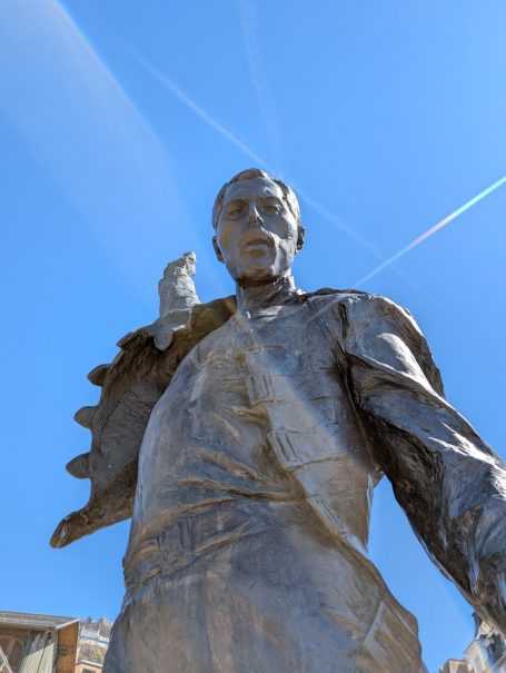Bronze statue of a Freddie Mercury holding a microphone, set against a clear blue sky.
