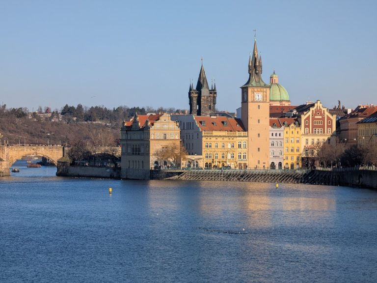 Historic buildings along a river, with a bridge in the background and clear blue sky.