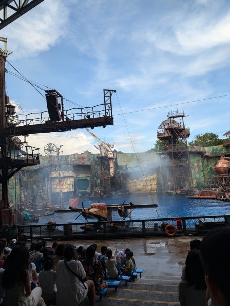 A large outdoor water stage with spectators, industrial-themed set, and blue water.