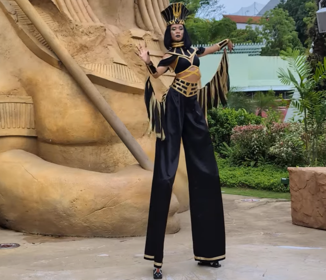 A performer in an elaborate costume poses on tall stilts near a large statue.