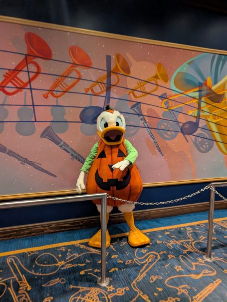 A character in a pumpkin costume poses in front of a colourful musical instruments mural.