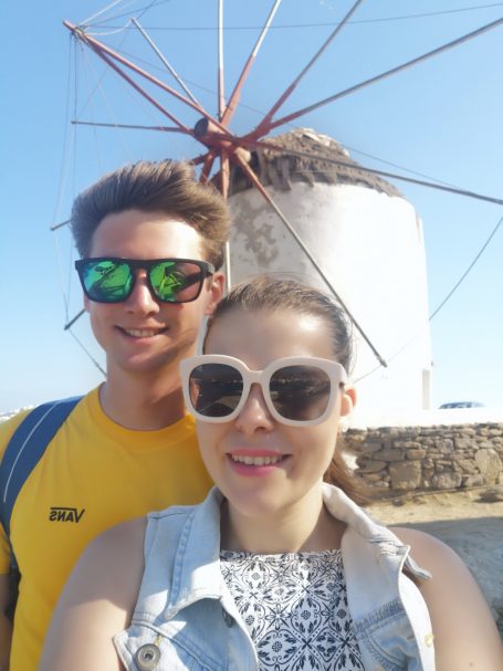 I&B's Founder and Husband posing in front of the three traditional Windmills of Mykonos with thatched roofs against a clear blue sky.