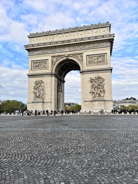 The Arc de Triomphe stands tall against a cloudy sky, surrounded by a bustling plaza.