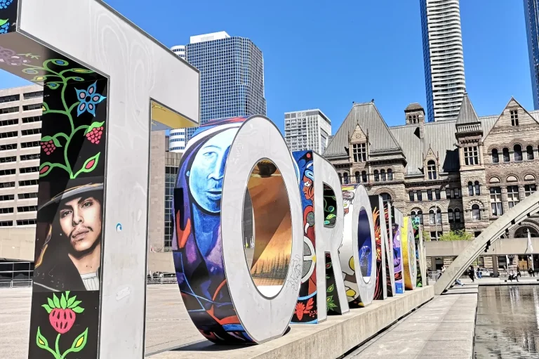 Colourful "Toronto" sign in front of modern buildings and historical architecture in Nathan Phillips Square.