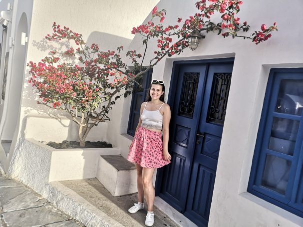 I&B's Founder posing in front of a blue door with pink flowers surrounding in the background. Located in a cobbled street in Oia, Santorini