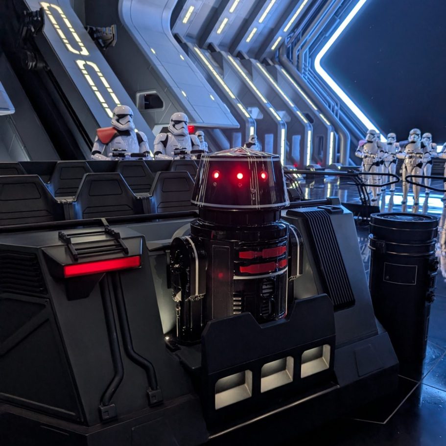 A dark robot with red eyes stands in a futuristic setting with stormtroopers in the background.