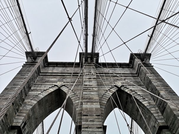 View of the Brooklyn Bridge's arches and cables from below, against a grey sky.