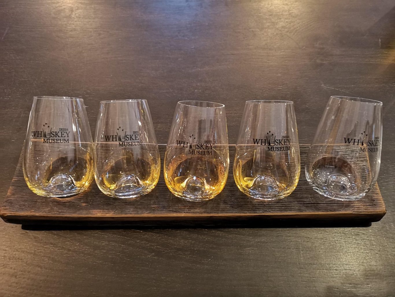 Five transparent glasses filled with yellow liquid on a wooden tray.