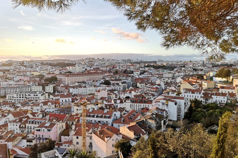 Panoramic view of a Lisbon with red-roofed buildings and distant mountains under a blue sky.