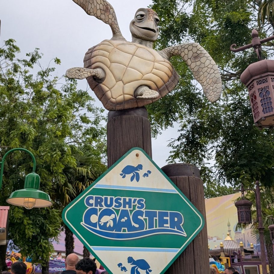 Sign for "Crush's Coaster" with a turtle figure above it, set in a lively amusement park.