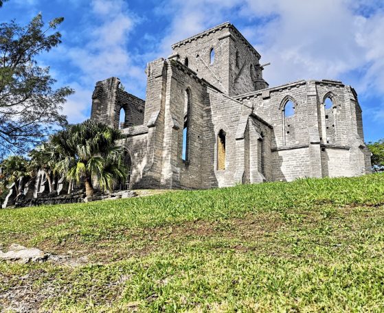 The Unfinished Church, a ruined stone building with arched windows, surrounded by grass and trees against a blue sky. Located in Bermuda.