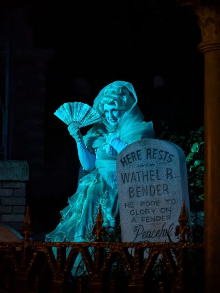 A ghostly figure in a blue hue holds a fan beside a gravestone at night.
