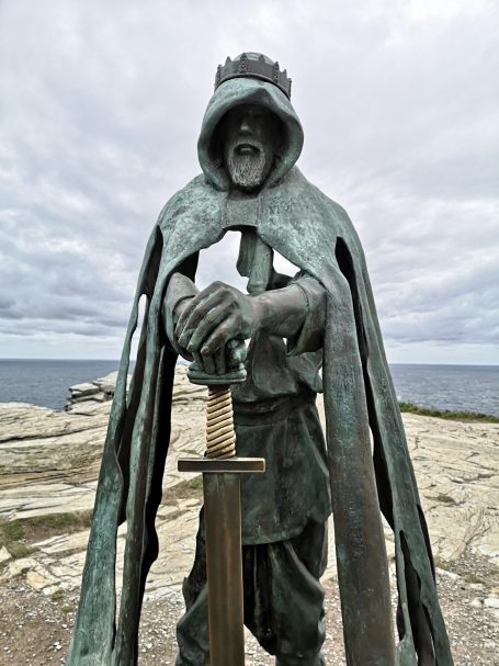 Bronze statue of a hooded figure holding a sword, facing the sea under cloudy skies.