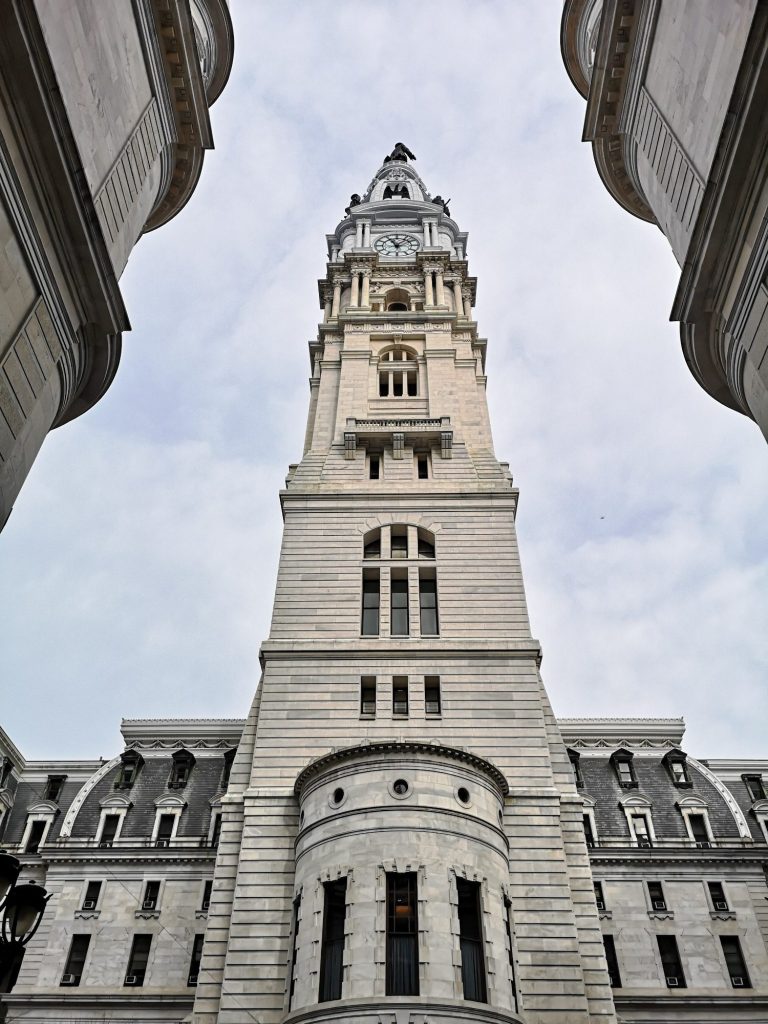 View of the tall historic City Hall building with an ornate clock tower against a cloudy sky, located in Philadelphia. 