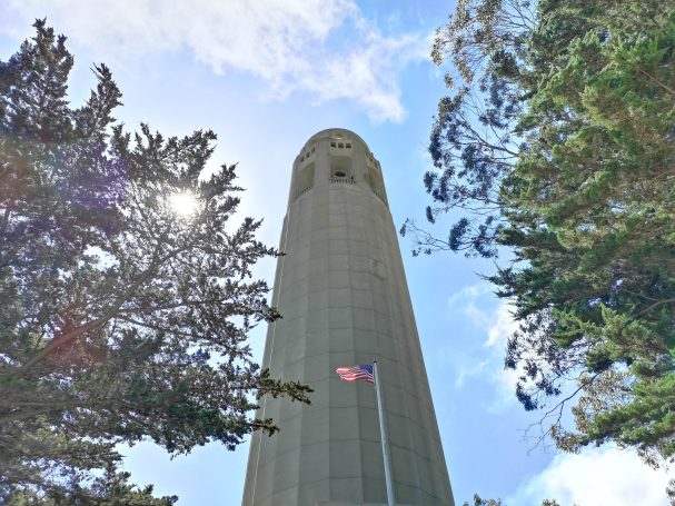 Coit Tower rises above trees, with the American flag visible at its base against a blue sky.
