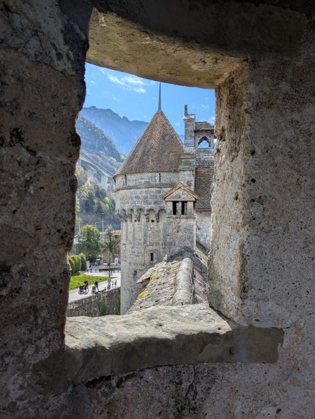 View of Chillon Castle tower through a stone window, framed by rugged walls and blue sky.