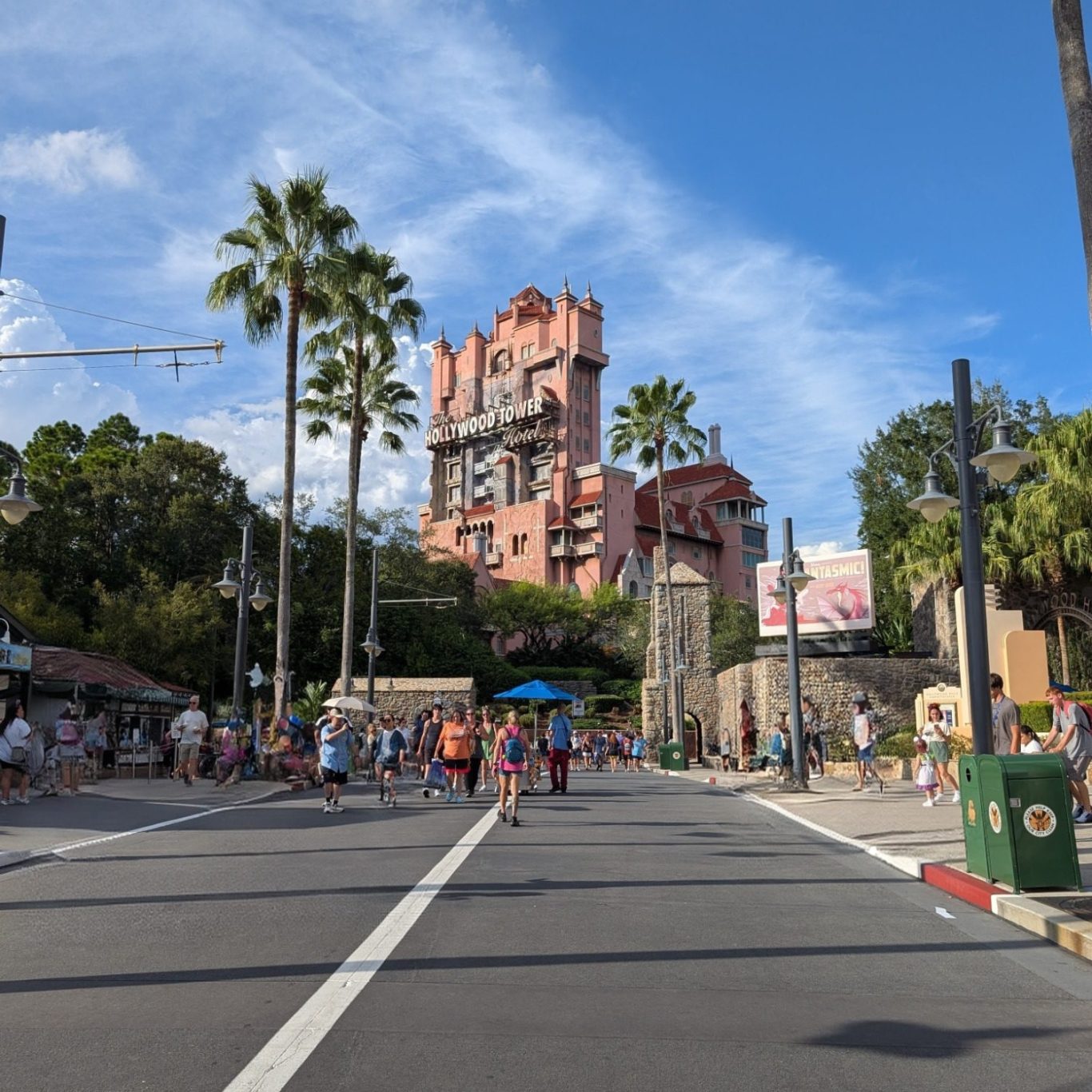 View of a theme park street leading to a tall, iconic hotel tower surrounded by palm trees.