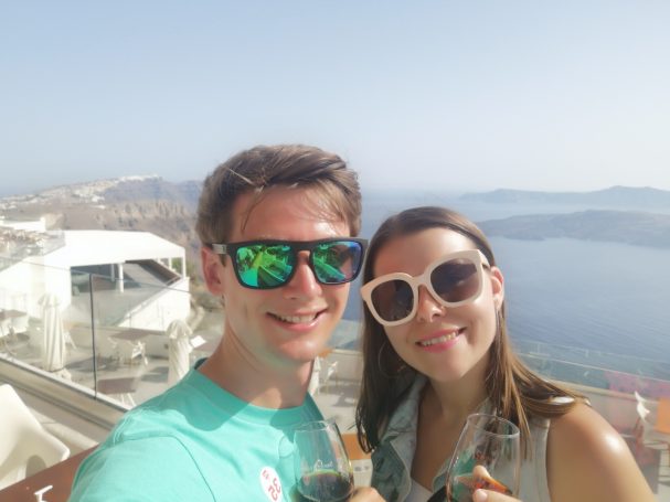 I&B's Founder & Husband posing with wine glasses at Santos Wines with the Santorini coastline in the background.