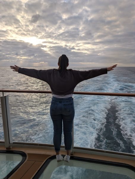 I&B's Founder posing in a 'T' shape on the back of a cruise ship, standing on a glass walkway with the ocean and sunset in the background.