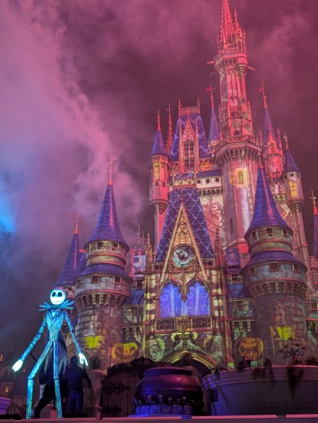 A spooky character stands in front of a brightly lit castle under swirling clouds.