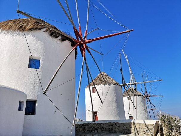 Three traditional Windmills of Mykonos with thatched roofs against a clear blue sky.