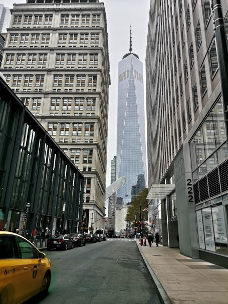 View down a New York city street towards the One World Trade Center, with a yellow taxi on the road.