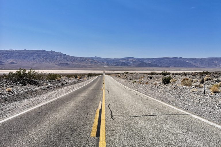 A long, straight road in Death Valley National Park, stretches into the distance amidst a desert landscape under a clear blue sky.