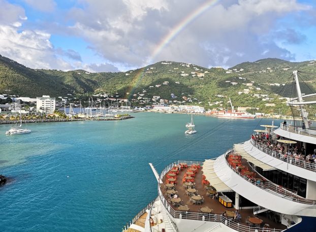 A cruise ship docked near a harbour with a rainbow arching over green hills.