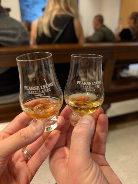 Two small glasses of whisky held in hands, with people blurred in the background.