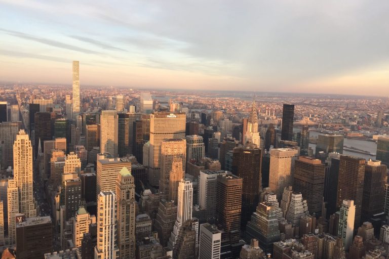 Aerial view NYC from the top of the Empire State Building, with a sprawling cityscape at sunset, with skyscrapers and distant horizons.