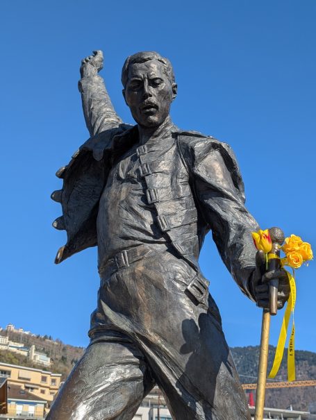 Statue of Freddie Mercury with a raised arm, holding flowers, against a blue sky.