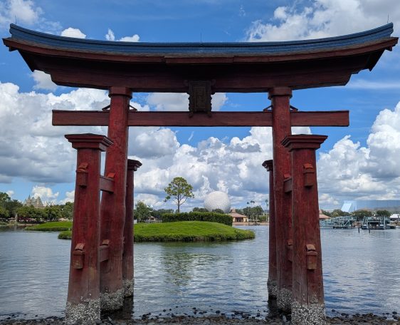 Red torii gate on the water with greenery and cloudy blue sky in the background.