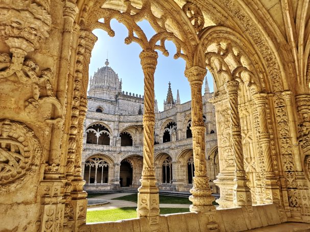 Gothic arches frame a courtyard with ornate stone detailing and historical architecture, located at Jeronimos Monastery in Lisbon.