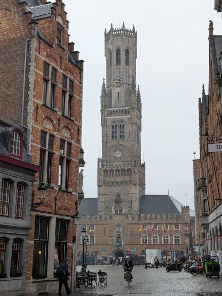 Tall belfry tower amid historic buildings in a rainy, cobbled street scene.