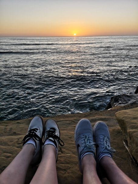 Two pairs of sneakers are resting on the rocky shore of the Sunset Cliffs at sunset, with ocean waves in the background.
