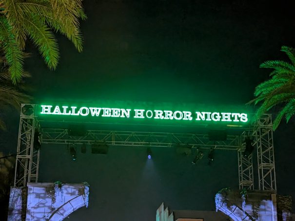 Neon green sign reading "Halloween Horror Nights" under a night sky with palm trees.