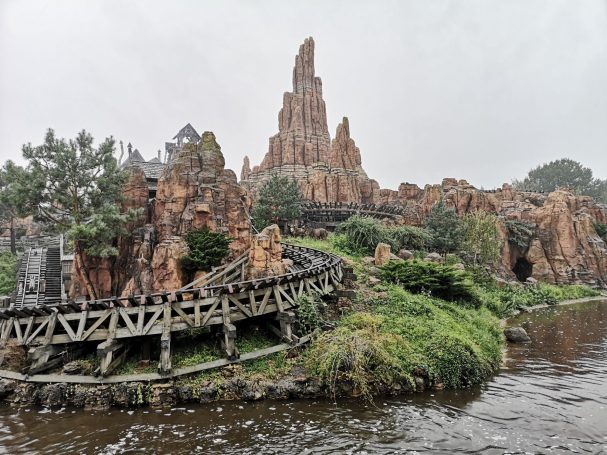 Rocky landscape featuring a wooden roller coaster beside a river, under a cloudy sky.