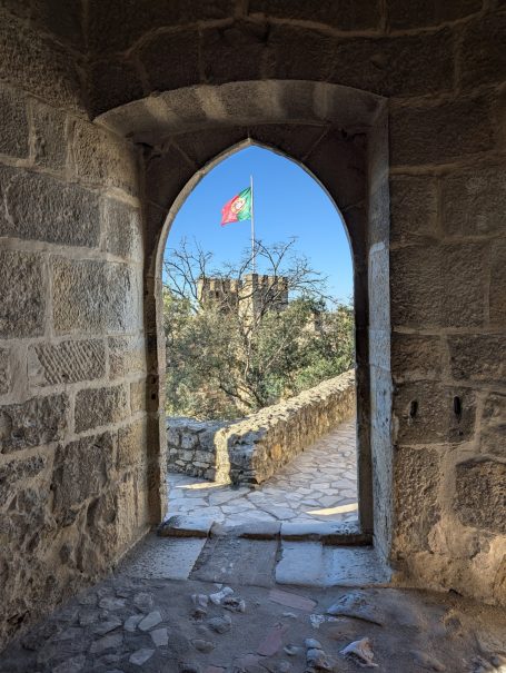 View of Sao Jorge Castle in Lisbon through a stone archway featuring the castle walls and Portuguese flag against a clear sky.