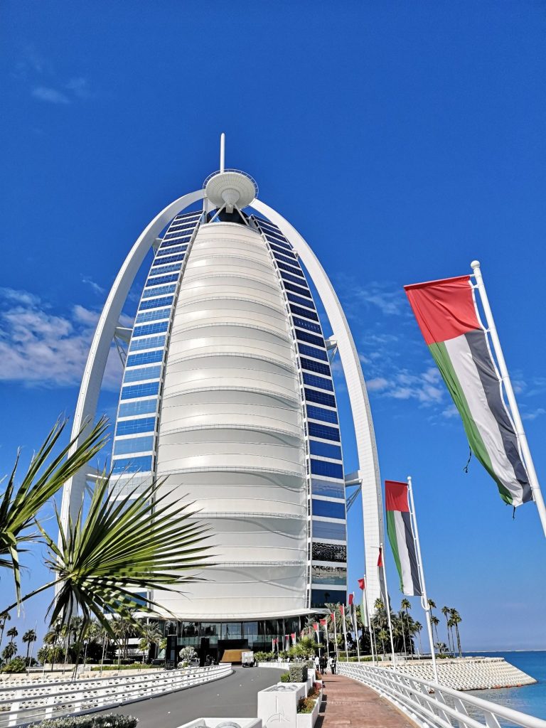 Burj Al Arab tower with palm trees and flags against a clear blue sky.
