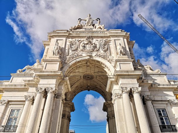 View of the Rua Augusta Arch, a white stone archway with sculptures, against a blue sky with scattered clouds.