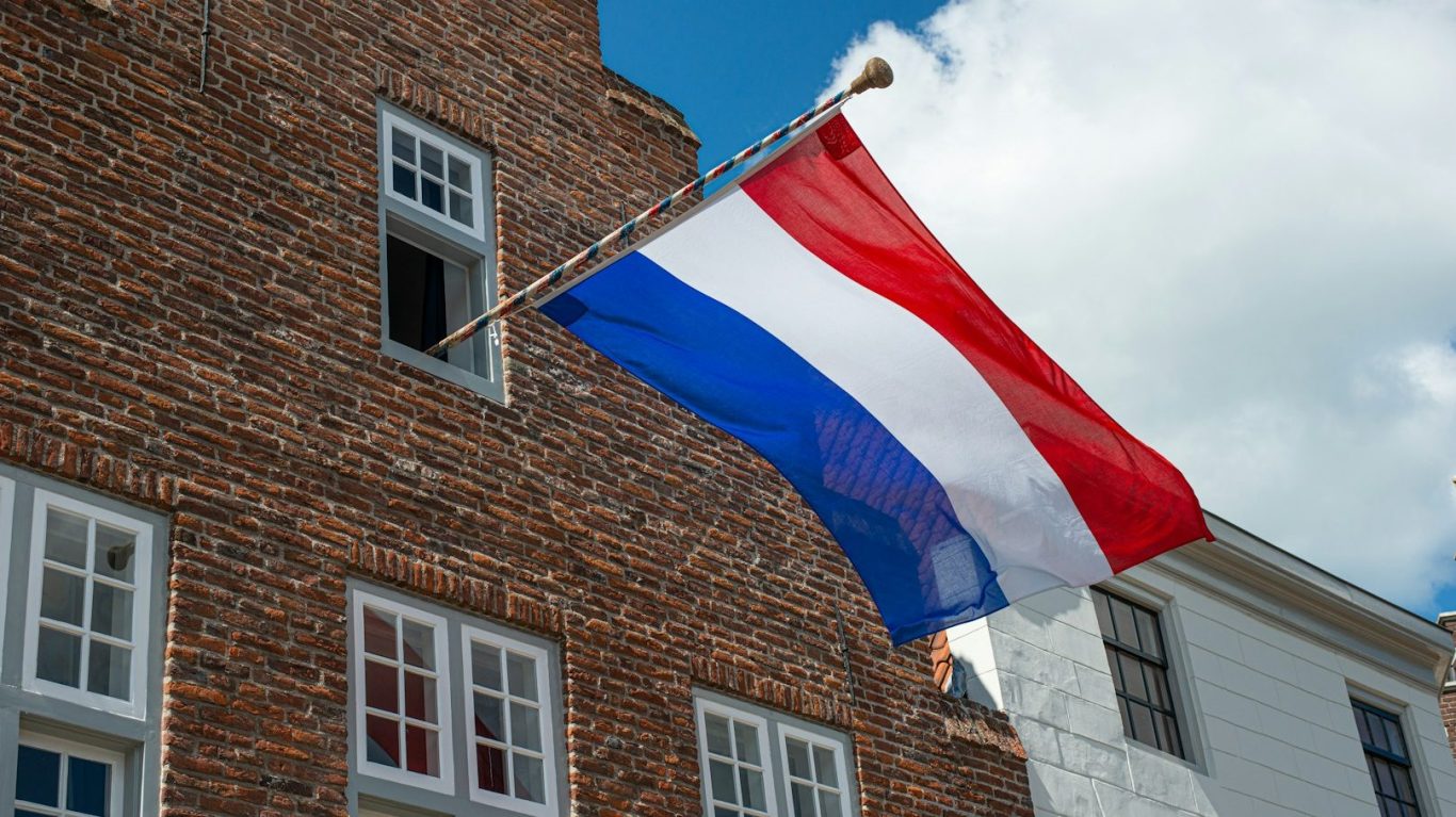Dutch flag waving from a window of a brick building under a cloudy sky.