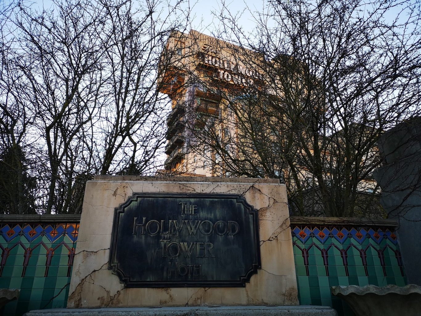 A weathered plaque in front of a tall, partially obscured building and bare trees.