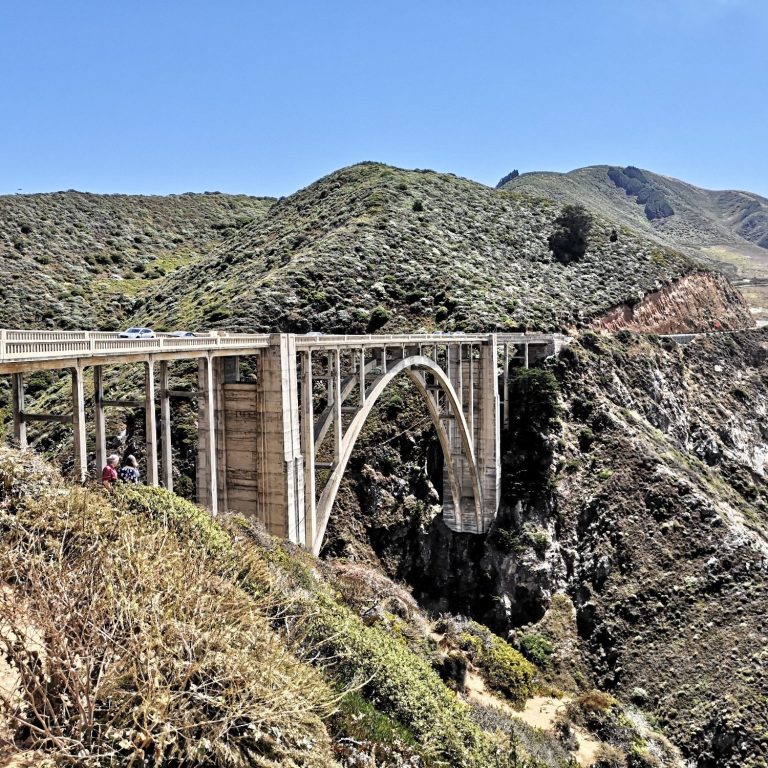 A large arch bridge, known as Bixby Creek Bridge spans a rocky canyon with green hills in the background.