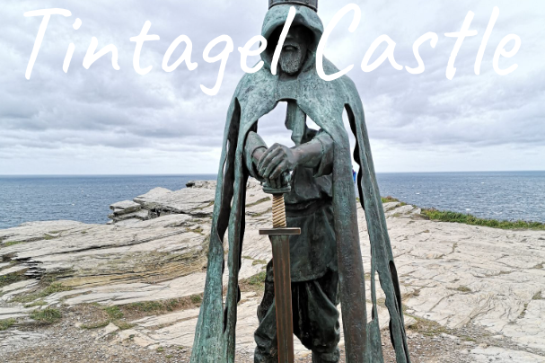 Statue of a cloaked figure holding a sword near Tintagel Castle by the sea.