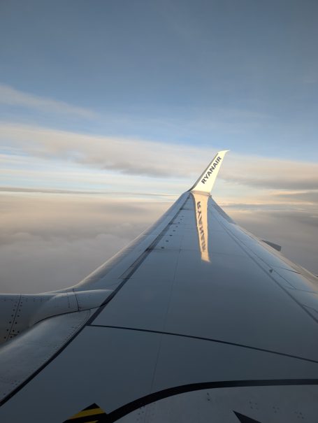 Wing of a Ryanair airplane with a clear sky and fluffy clouds visible in the background.