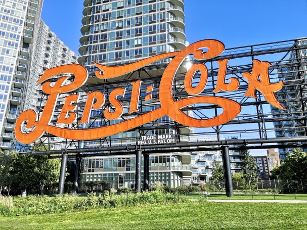 Bright orange retro Pepsi-Cola sign in an urban park setting with buildings in the background.