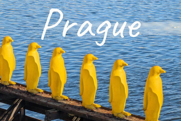 Seven yellow penguins standing on a wooden pier by water, with the word "Prague" above.