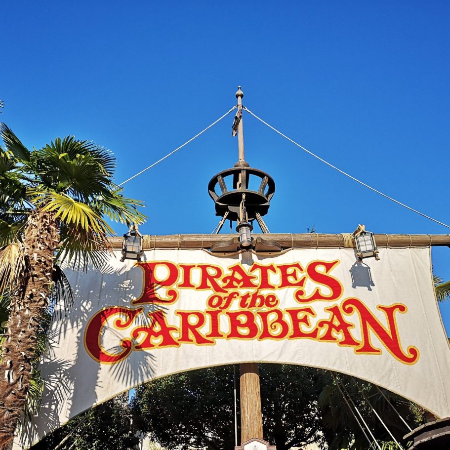 Sign for "Pirates of the Caribbean" featuring tropical foliage and a clear blue sky.